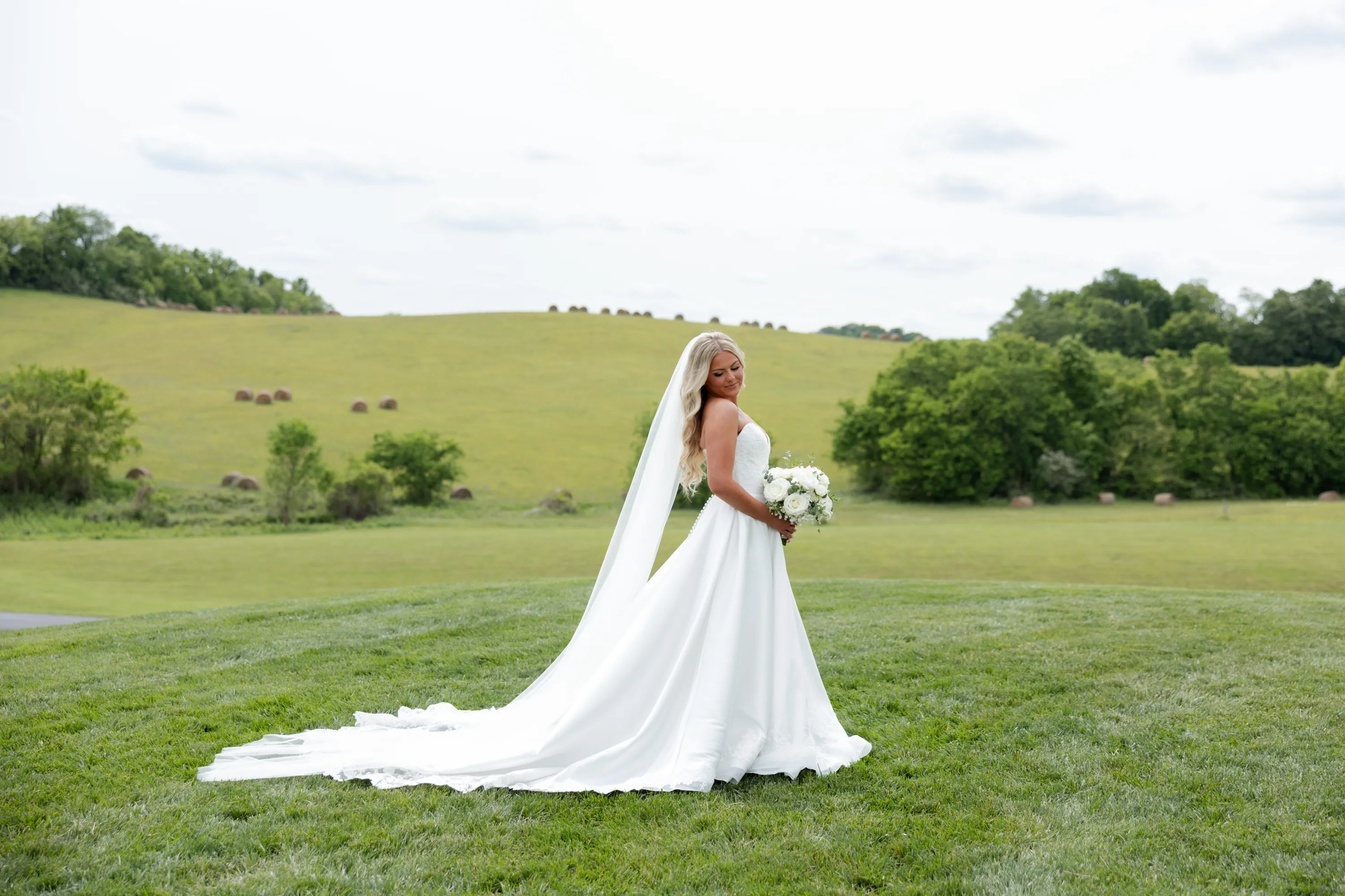 Taylor full-length in her gown and veil, hay bales on the hills behind her