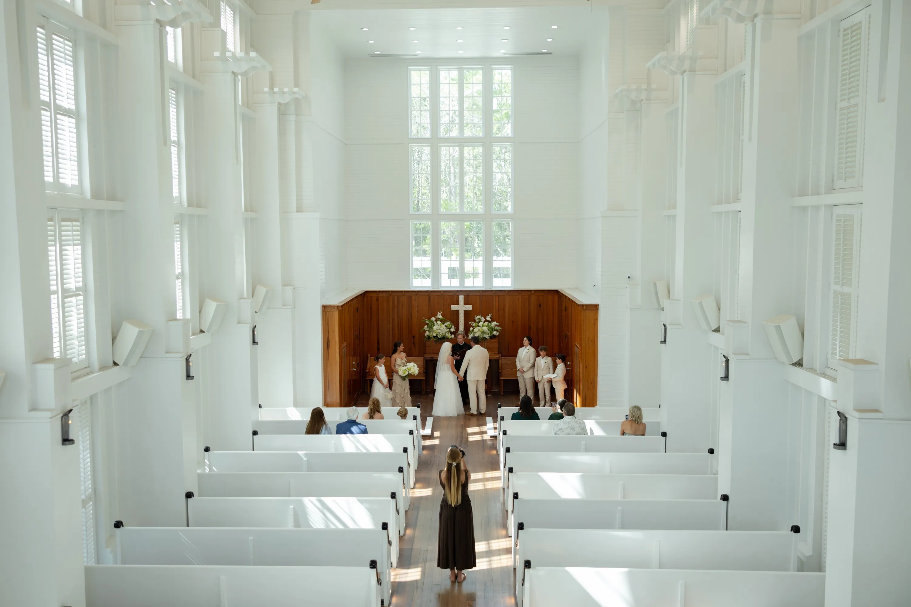 Brietyn Park photographing a wedding ceremony from the aisle of a white chapel, the couple at the altar ahead of her
