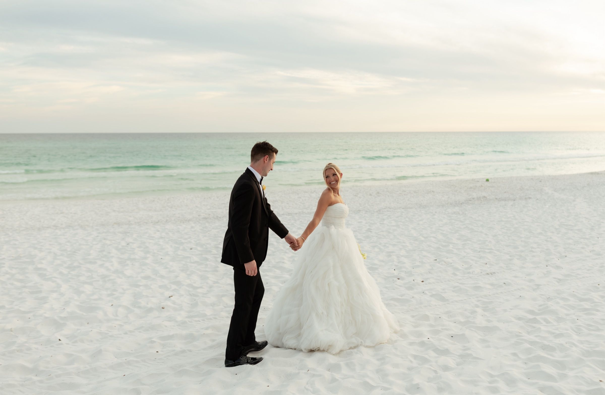 A couple hand in hand on white sand at sunset, the gulf soft and pale behind them