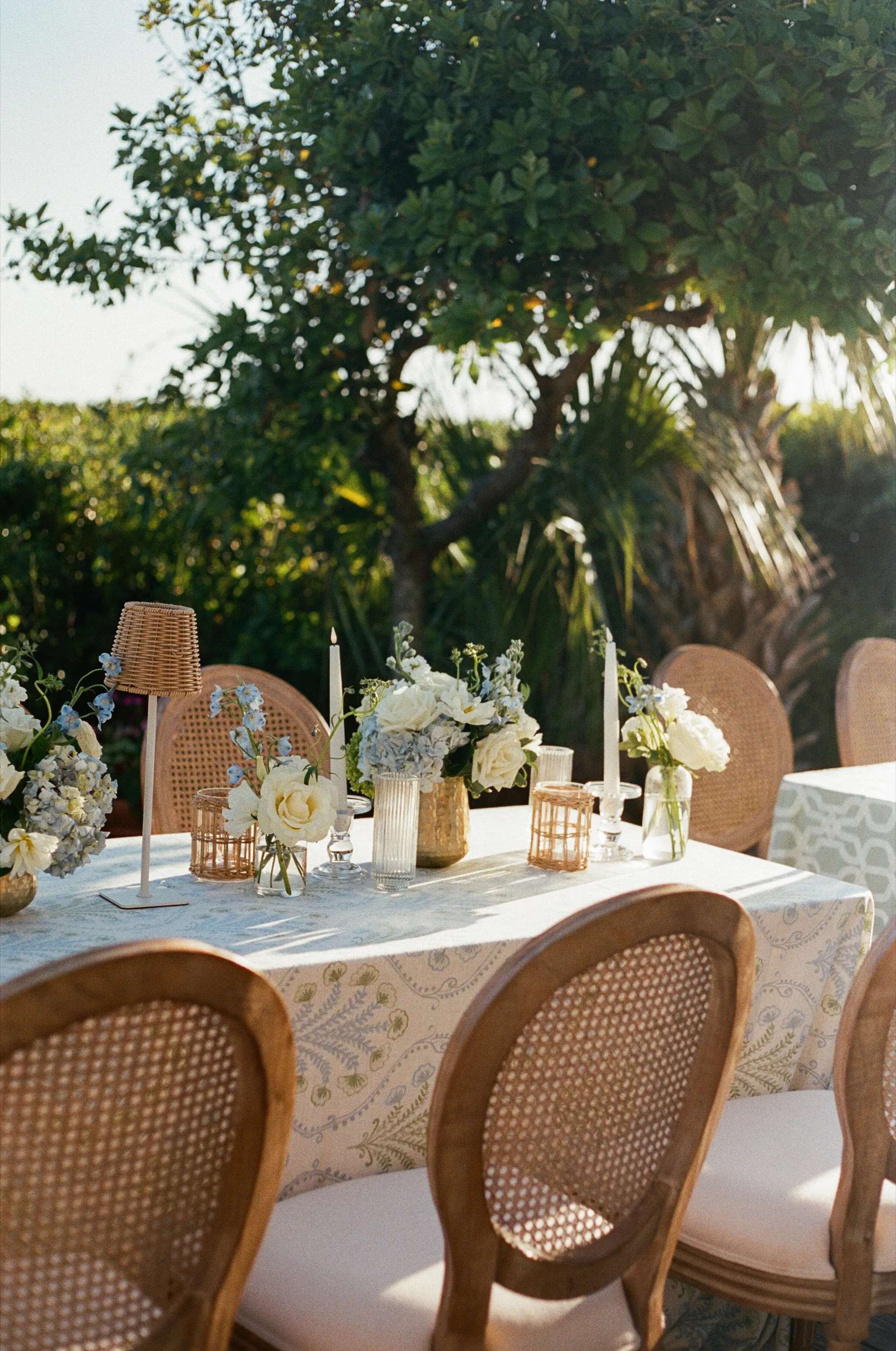 A reception table in the evening sun — cane-back chairs, taper candles, and blue-and-white garden flowers
