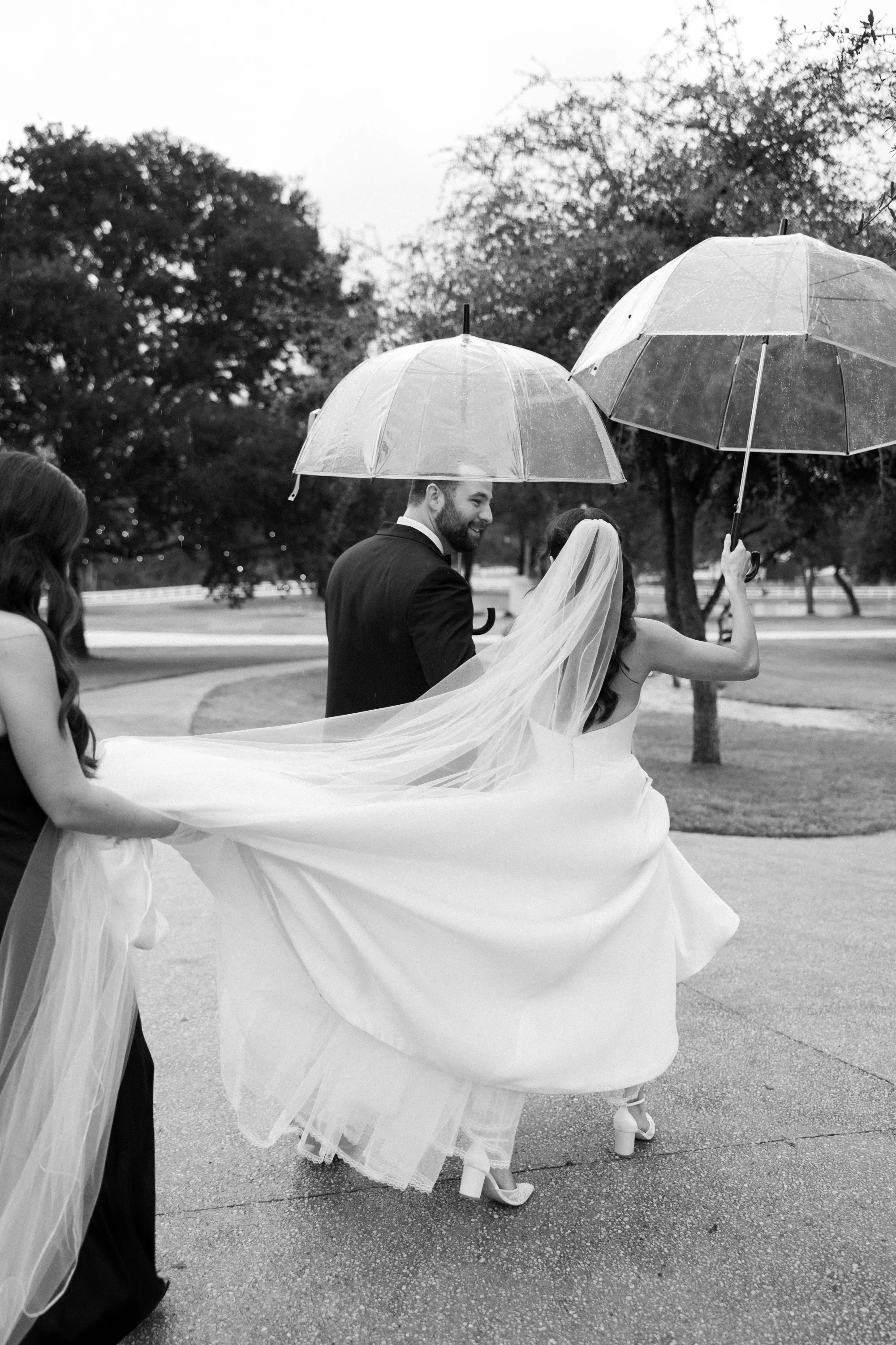 Walking through the rain under clear umbrellas, veil catching the wind, in black and white