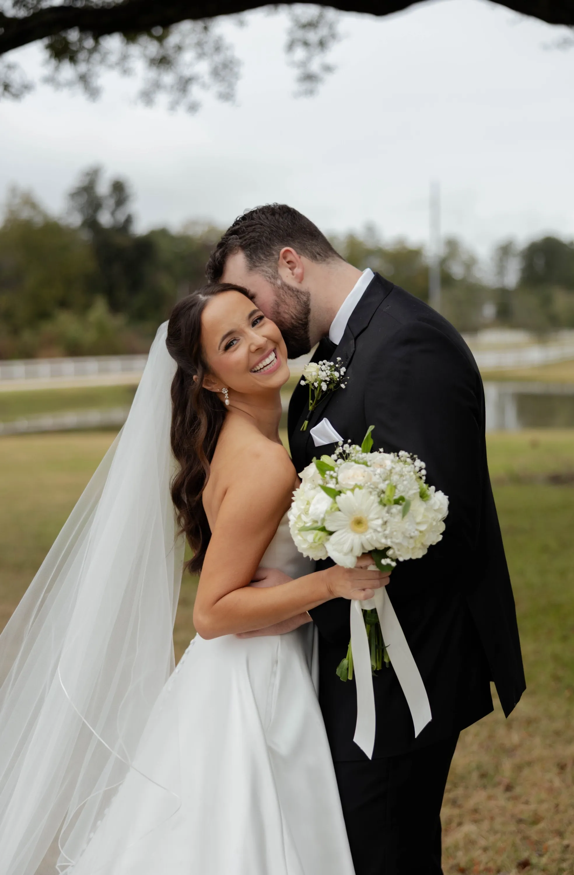 Shelbie beaming as Wilson kisses her temple, the pond behind them