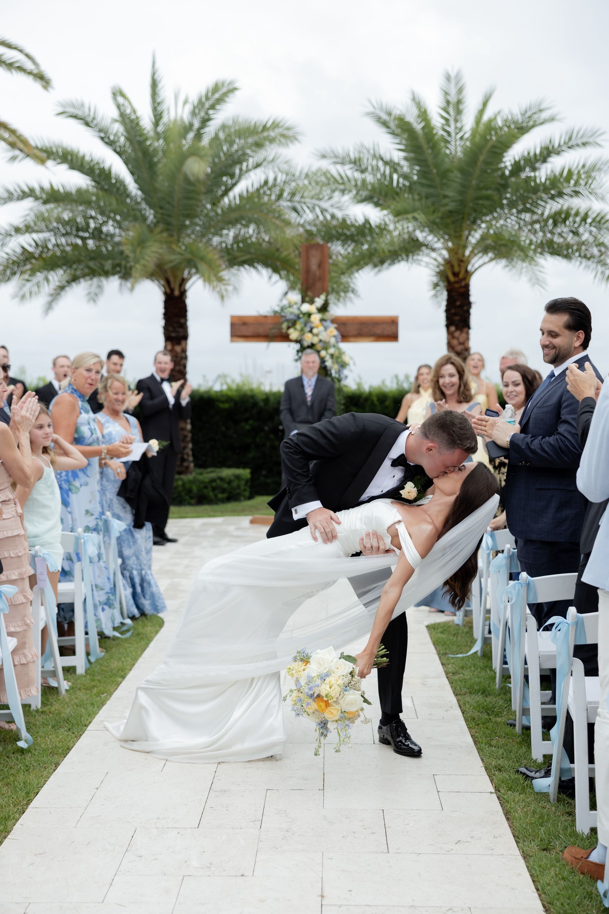 Will dips Savannah into a kiss on the white stone aisle at The Alice, palms and a flower-dressed cross behind them