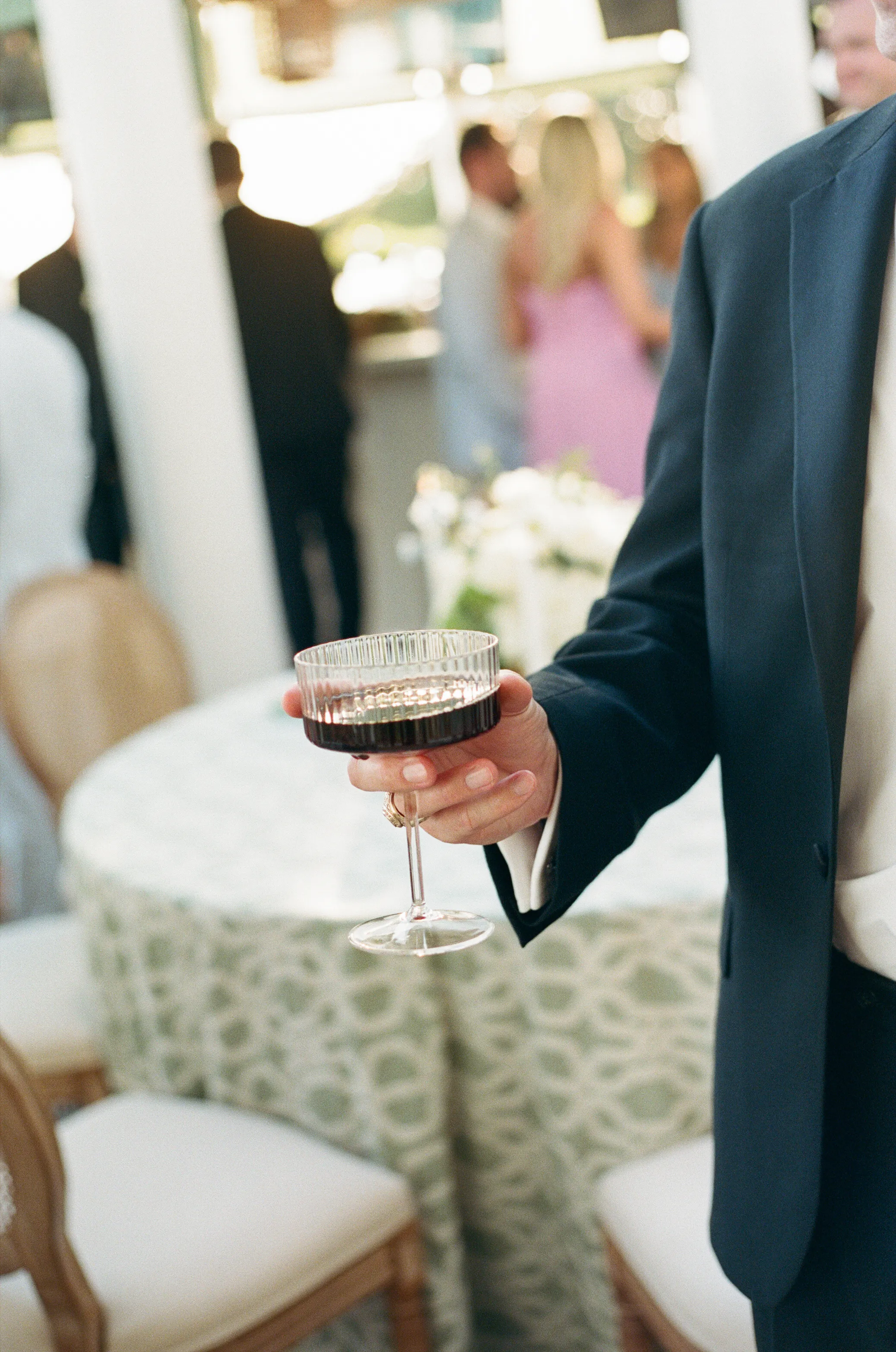 A guest's hand holding a coupe glass at an outdoor reception, patterned linen in the background