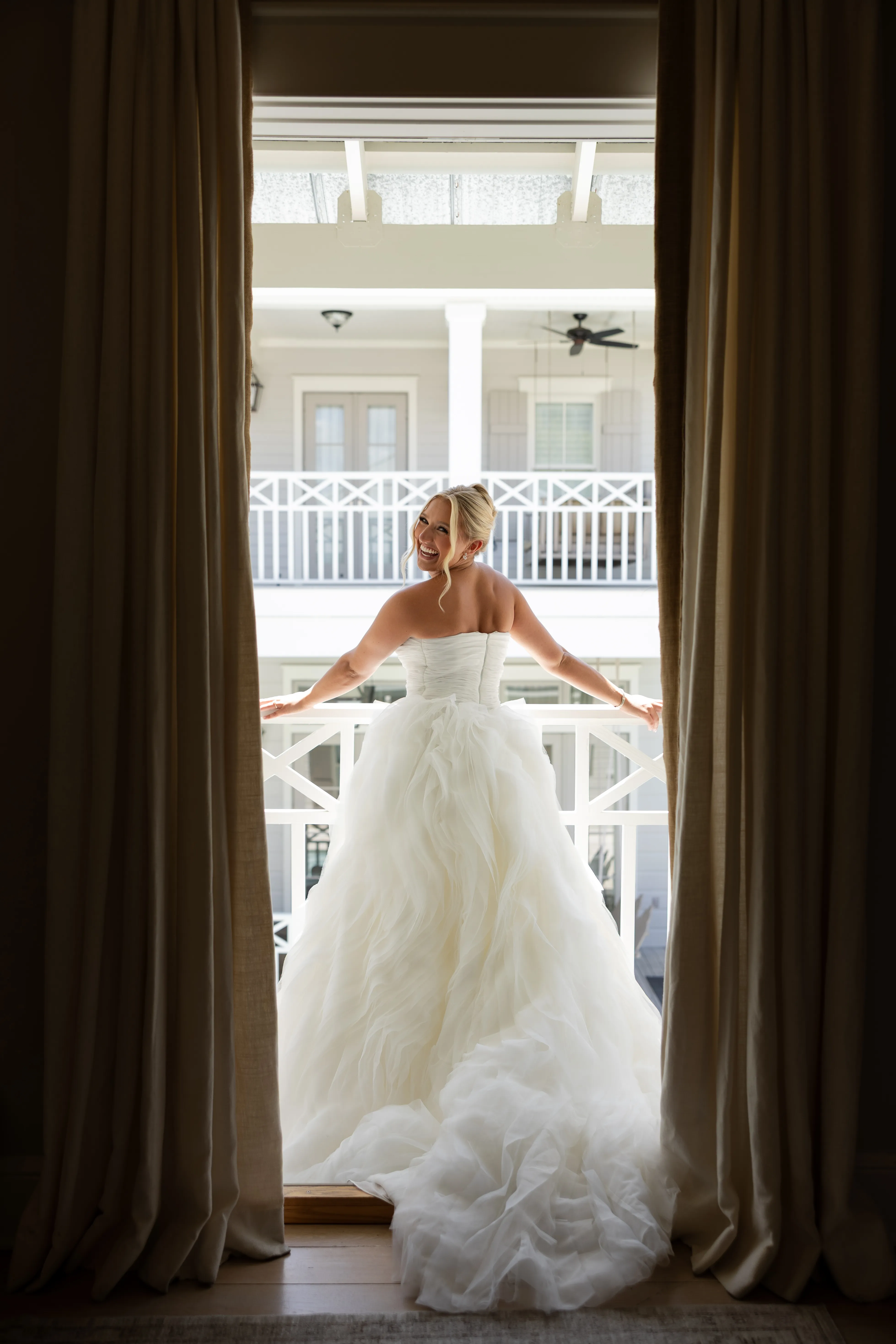 A bride seen from behind, framed by tall curtained windows as she steps toward a balcony