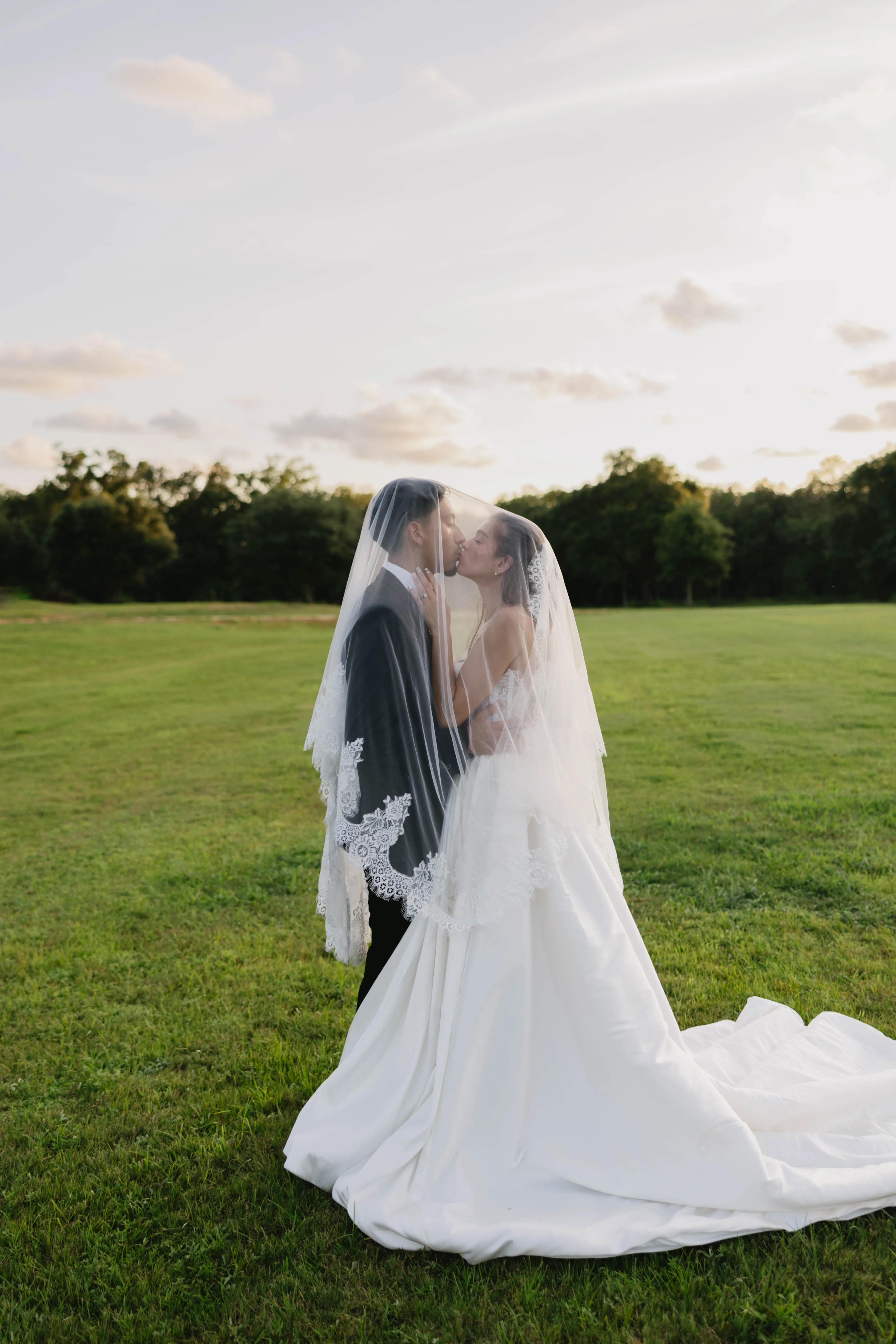 A couple kissing beneath the bride's veil in an open field at sunset