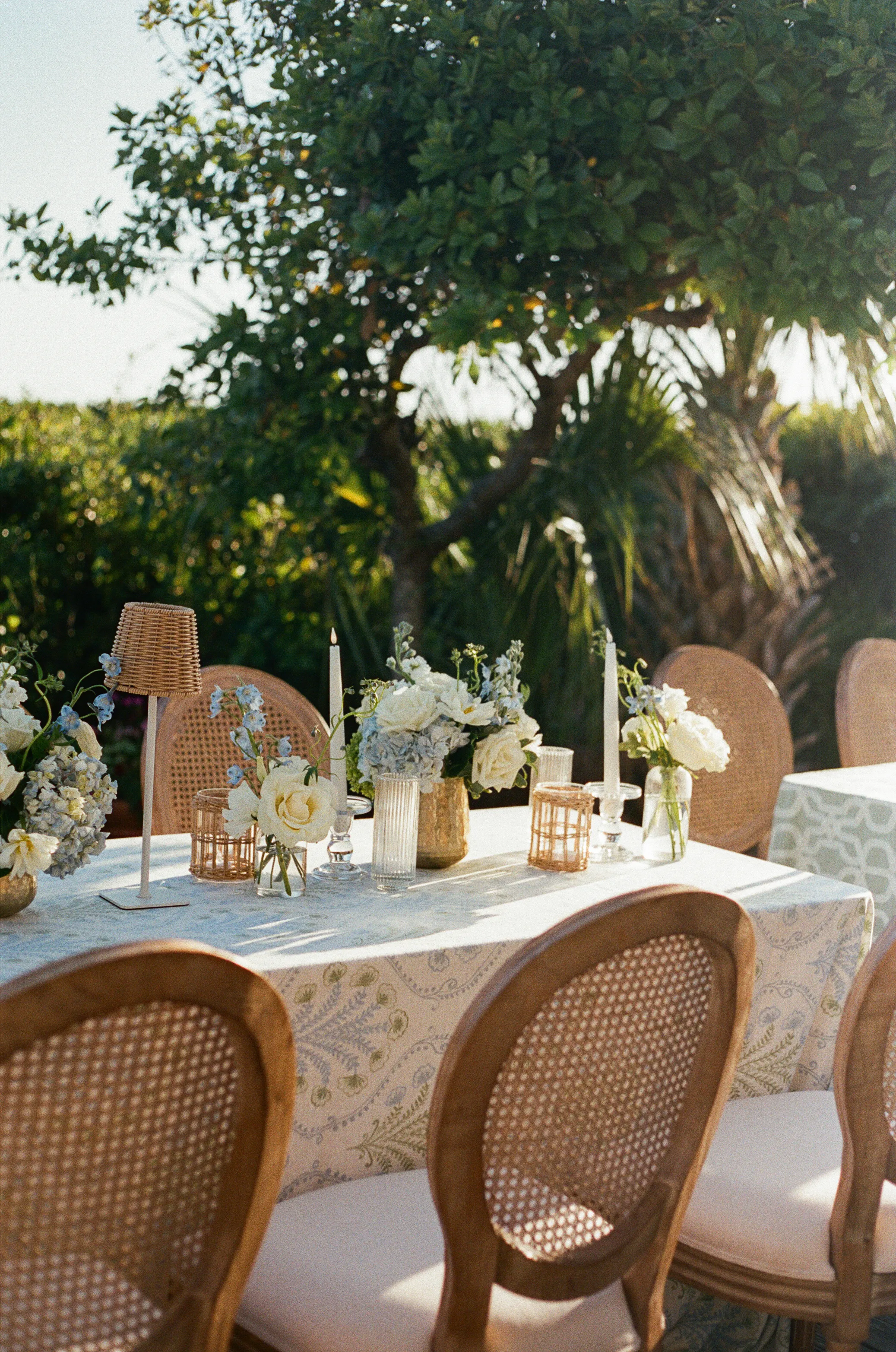A reception table set with woven chairs, patterned linen, and low arrangements of garden flowers
