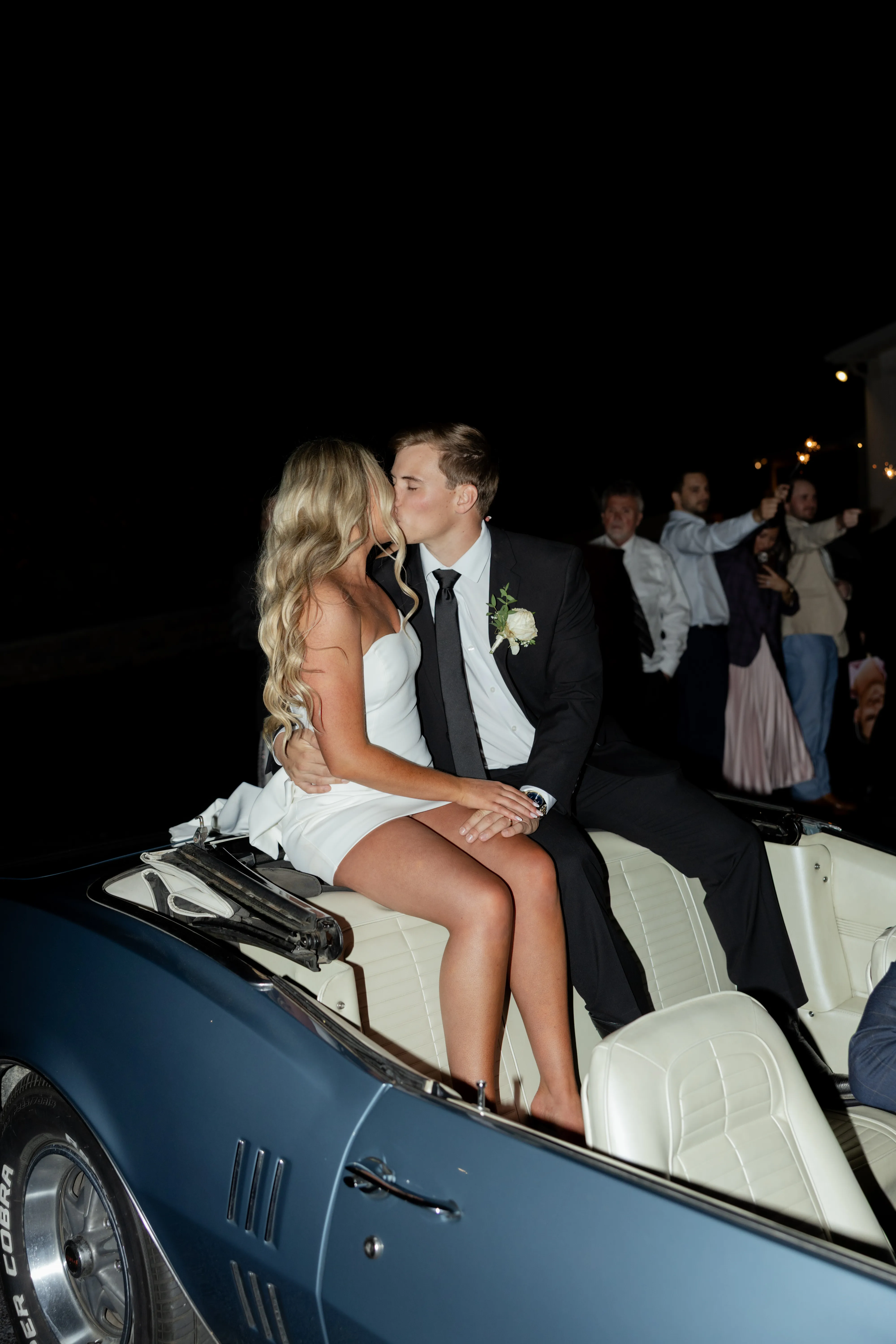 A couple perched on the back of a vintage convertible at night, guests celebrating around them