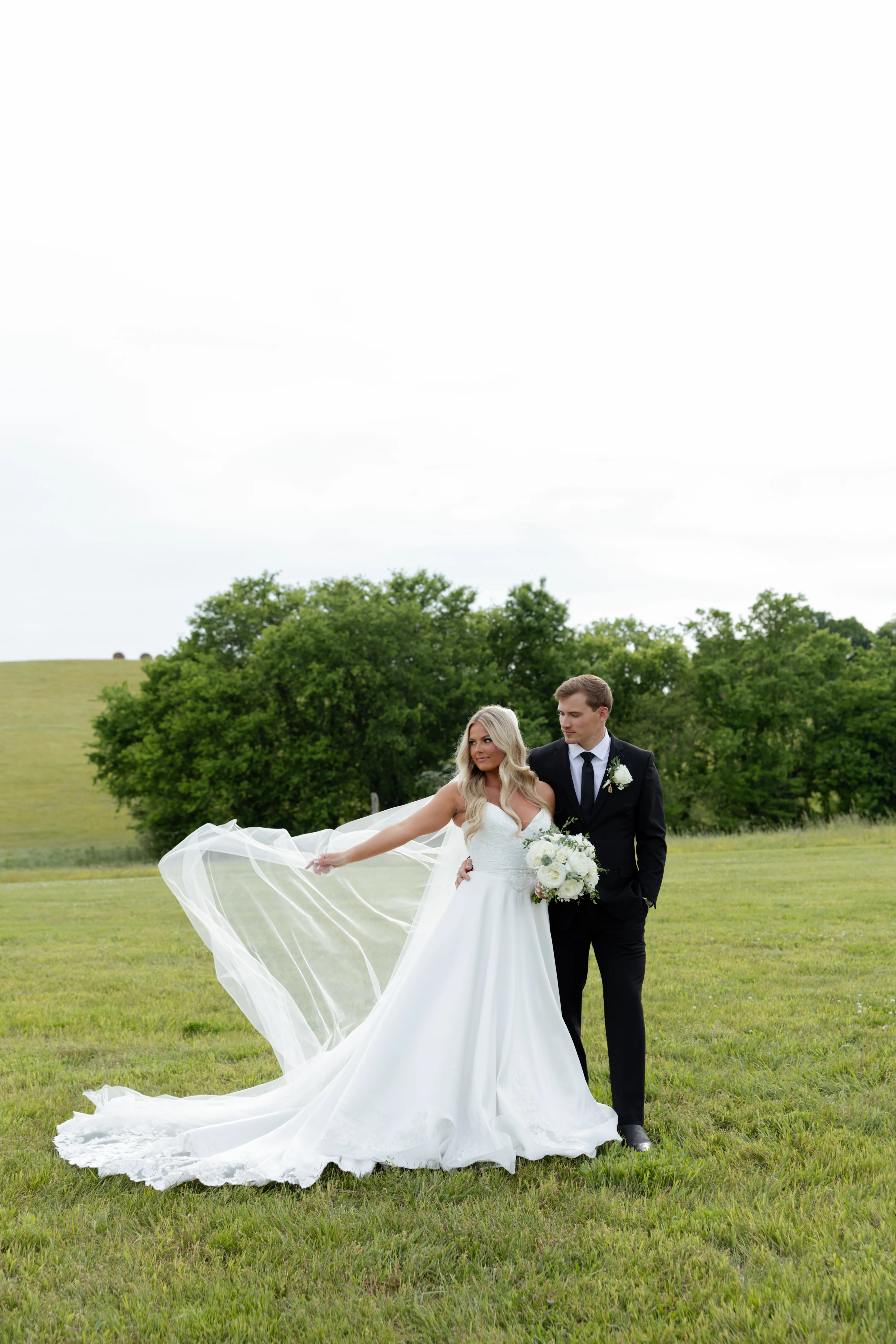 A couple standing together in a hillside field at golden hour, the bride's veil caught in the wind
