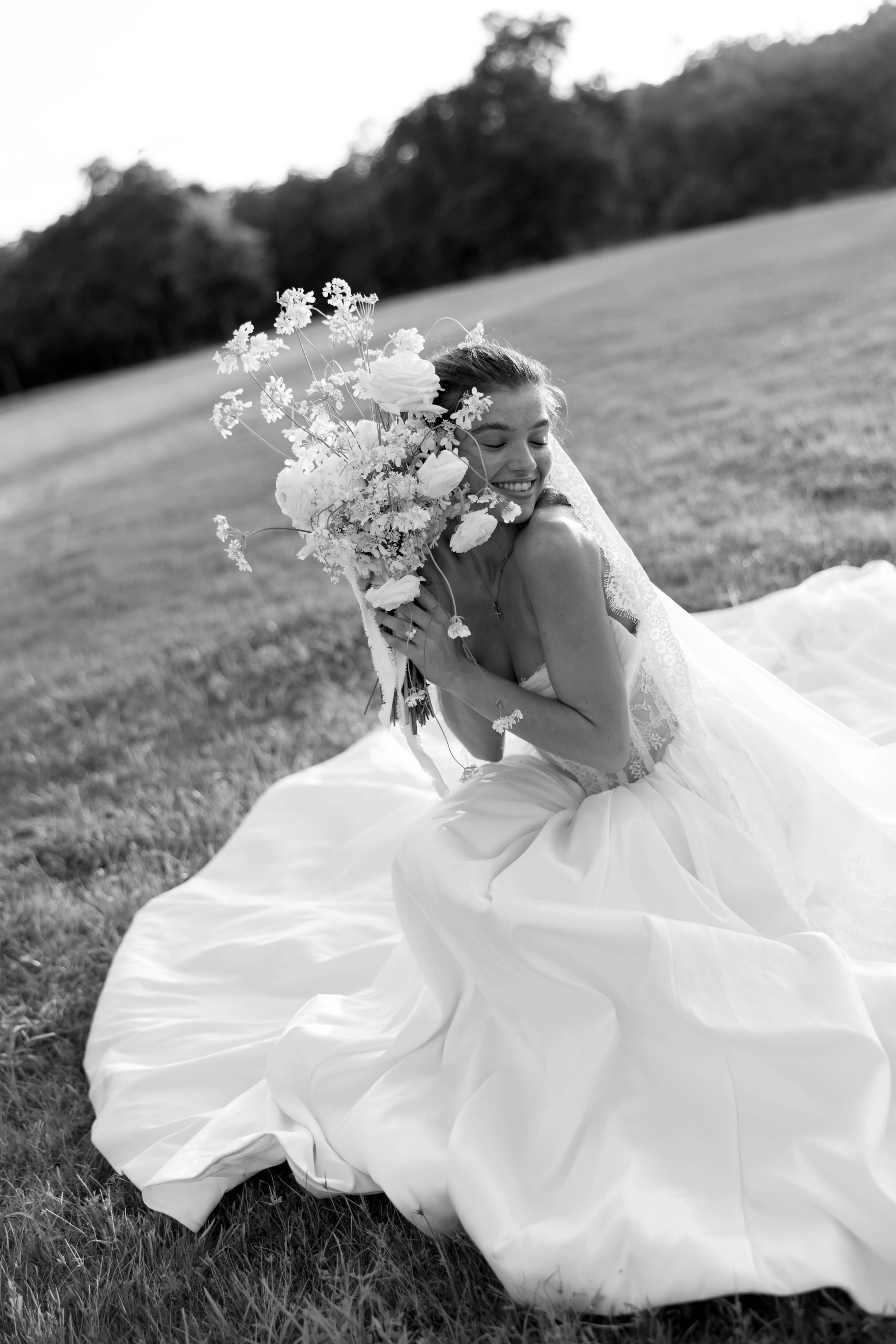 A bride seated in an open field, lifting her bouquet overhead, photographed in black and white