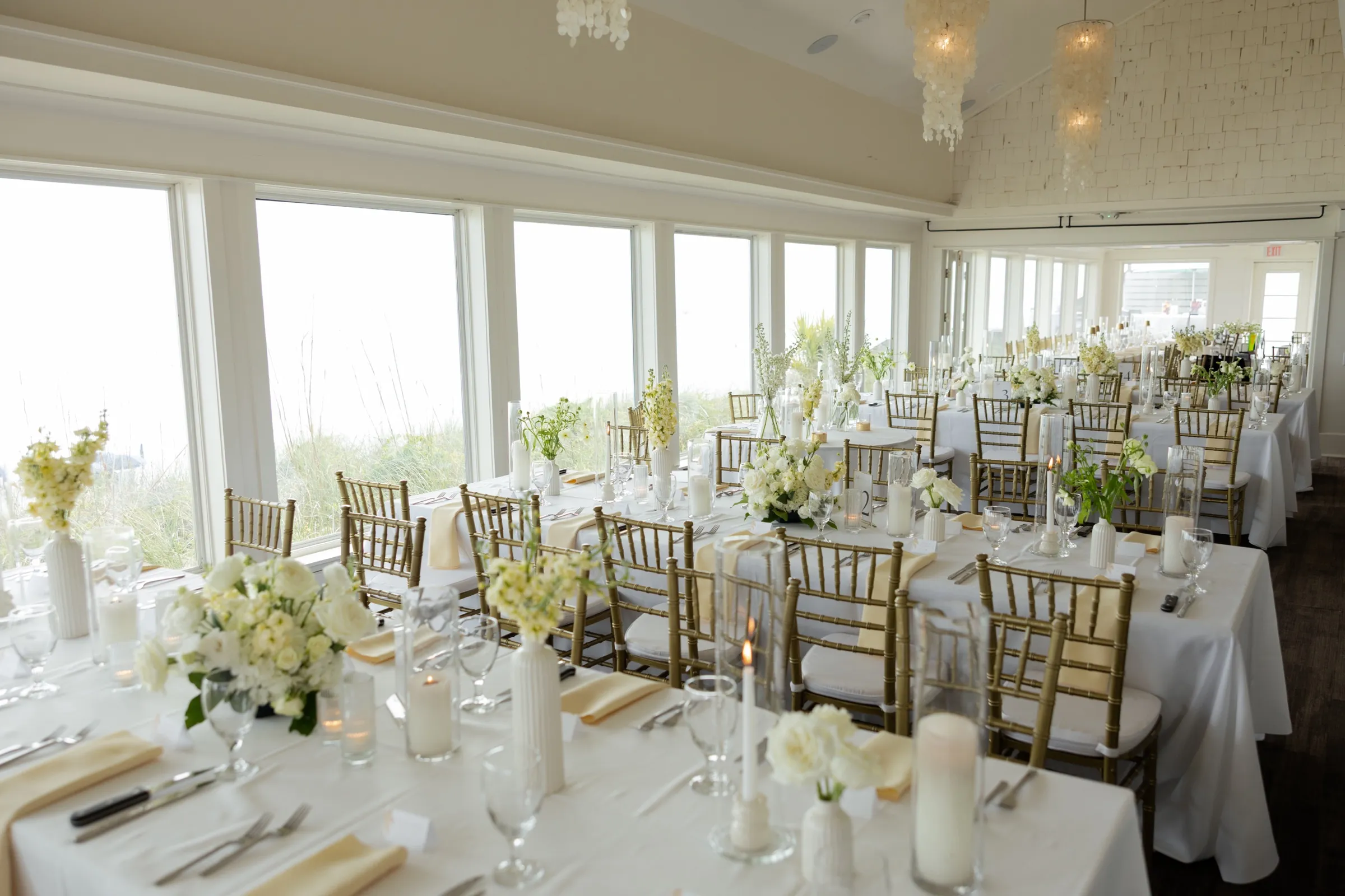 A reception room set for dinner in window light — white linens, gold chairs, and candles waiting for evening