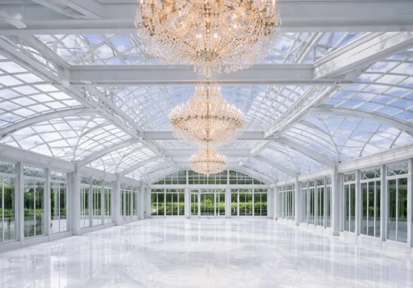 Elegant glass conservatory ballroom with crystal chandeliers, white floors, and lush garden views.